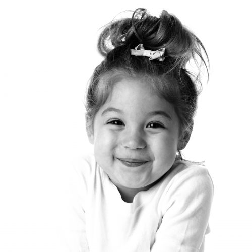 A head and shoulders image of a smiling real caucasian little girl shrugging her shoulders and giggling. She has brown hair and brown eyes.